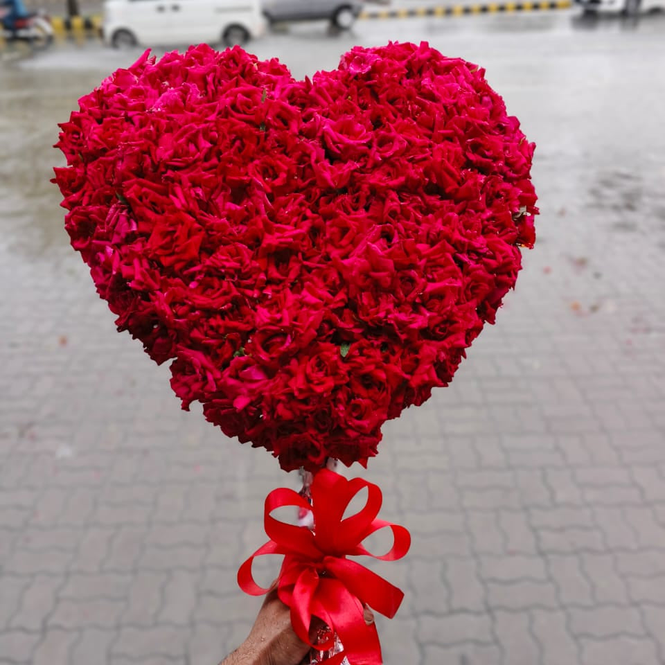 Heart-shaped bouquet of red flowers with a red bow held by a person on a street.