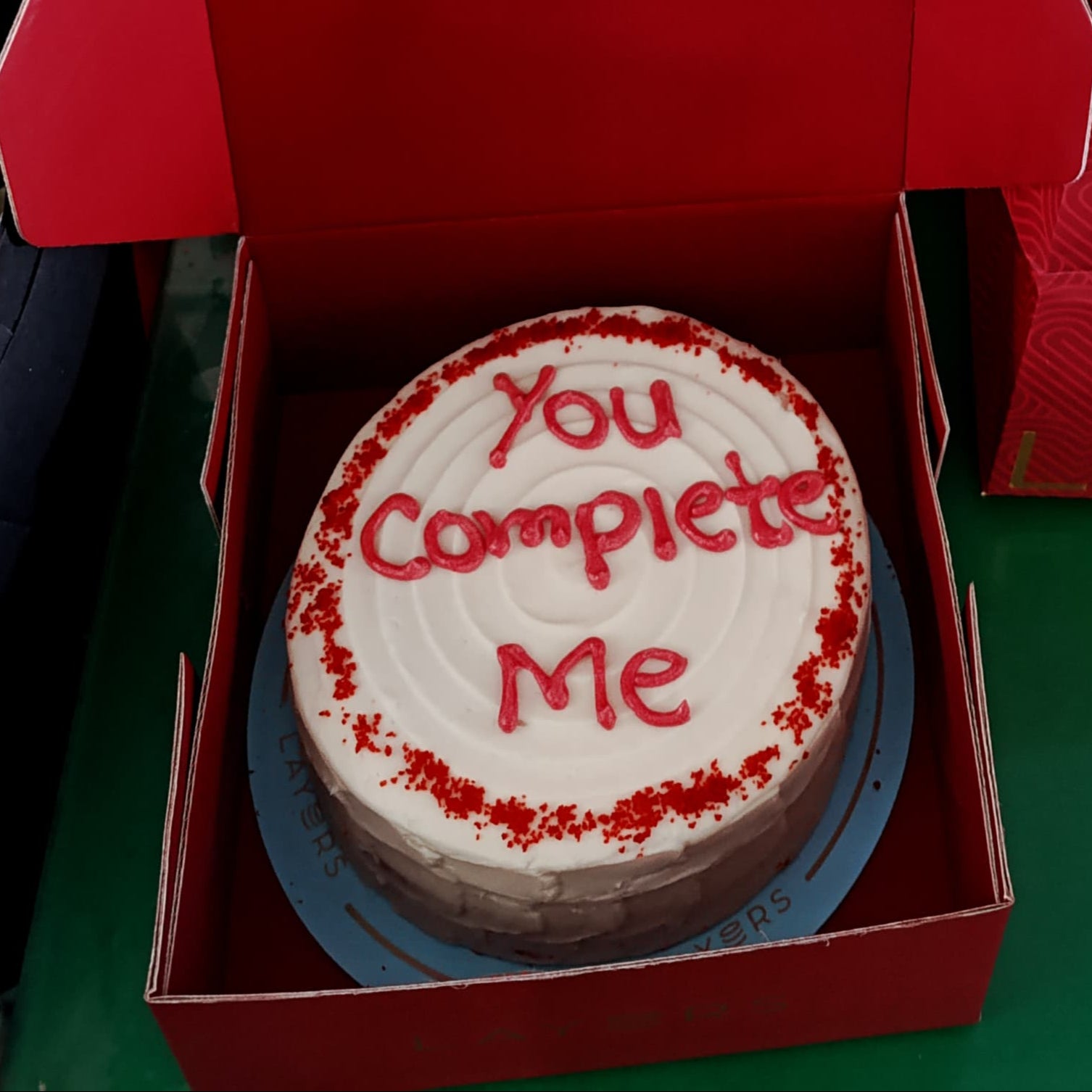 Bouquet of red roses and a cake with 'You Complete Me' text in front of more red roses.